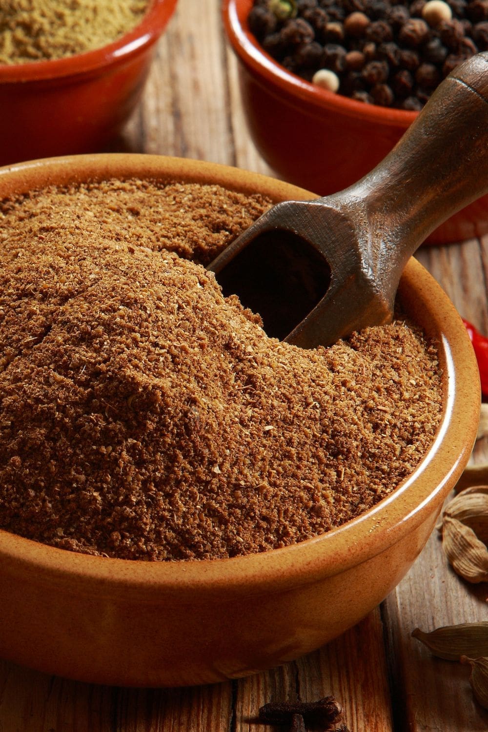 A bowl of garam masala powder with a wooden spoon on a dark wooden table.