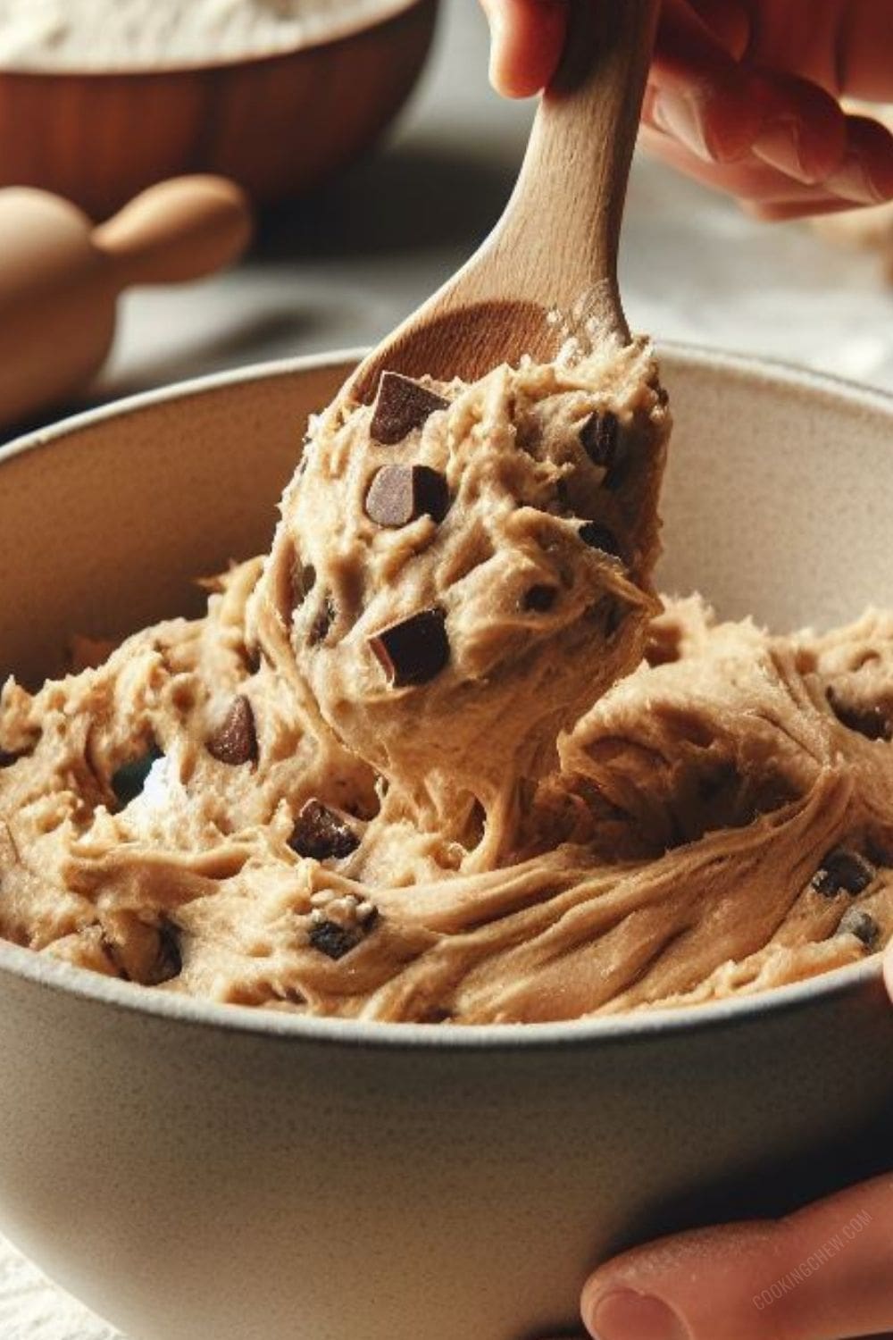 A hand mixing chocolate chip cookie dough with a wooden spoon in a stainless steel bowl.